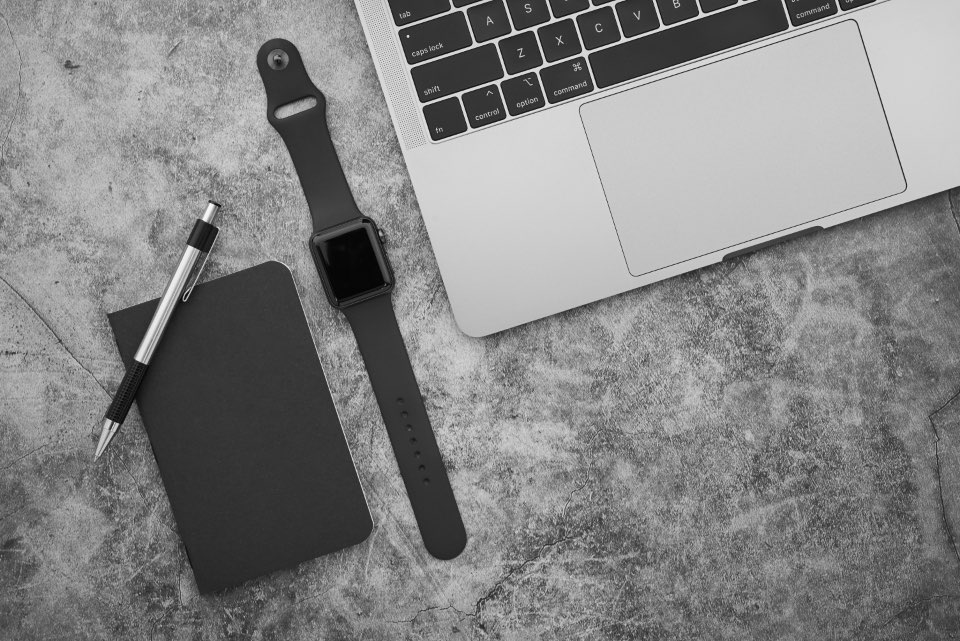 Caderno preto e caneta sobre mesa de concreto escura, ao lado de laptop e relógio